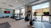 This fitness center at 1401 King in Bridgeport, Connecticut is a thoughtfully designed, single-floor facility built to support a full range of workout routines. The space is flooded with natural light through expansive floor-to-ceiling windows that line the walls, offering open outdoor views and creating an inviting, energizing atmosphere throughout. The interior features a neutral color palette with clean white walls, a teal accent wall, and a drop tile ceiling with recessed lighting that ensures consistent illumination across the entire floor.

The cardio zone is well-equipped with multiple treadmills, elliptical trainers, a stair climber, and a rowing machine, all positioned to take full advantage of the natural light and outdoor views. A dedicated strength training area features a multi-station cable machine, a flat utility bench, and a comprehensive rack of dumbbells spanning a wide weight range. Full-length mirrors are strategically placed to maximize the sense of space and support proper form during workouts.

Additional amenities include a wall-mounted flat-screen television, a built-in water bottle filling station and drinking fountain, and open floor space with exercise mats for stretching and functional training. The carpeted flooring and rubber mat zones provide comfort and safety throughout. The facility is clean, modern, and comprehensively outfitted for a high-quality fitness experience.