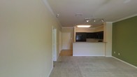 This single-floor residential unit offers an open-concept layout that connects the living area and kitchen in a cohesive, functional design. The living space is finished with plush wall-to-wall carpeting in a warm neutral tone, creating a comfortable and inviting atmosphere. Crown molding runs along the ceiling, adding a refined architectural detail that elevates the overall aesthetic.

The kitchen is positioned at the far end of the open space, featuring dark cabinetry that provides a striking contrast against the light-colored walls. A peninsula counter extends outward, offering additional prep space and serving as a natural divider between the kitchen and living area. Track lighting above the kitchen provides focused illumination, complementing the overhead flush-mount fixture. Black appliances complete the kitchen's contemporary look.

A hallway extends from the main living space, leading to additional rooms and the entry door, which is finished in classic white. The corridor features hard-surface flooring, transitioning from the carpeted living area. Neutral, light yellow-toned walls throughout the unit create a bright and airy ambiance, while the consistent use of white trim and molding ties the entire space together with a clean, polished finish.