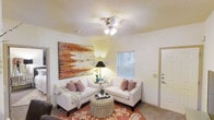 This single-floor space offers a warm and inviting atmosphere, thoughtfully designed for comfortable everyday living. The living area serves as the heart of the home, showcasing neutral wall tones that create a bright and airy feel. A ceiling fan with integrated lighting provides both functionality and a focal point overhead, while a textured ceiling adds a subtle architectural detail. Natural light filters in through a window fitted with blinds, contributing to the room's open and comfortable ambiance. The space features plush carpeting underfoot, complemented by a decorative area rug with bold, flowing patterns that adds a layer of visual interest. Seating consists of a pair of white upholstered sofas accented with patterned throw pillows, arranged around a stylish round ottoman that doubles as a coffee table. A large, vibrant abstract artwork in warm earth tones anchors one wall, serving as a striking decorative centerpiece. A side table with a classic table lamp adds a soft, ambient glow to the space. The layout is practical and well-proportioned, offering a seamless flow that makes the living area feel both functional and welcoming.