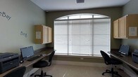 This single-floor space is designed as a practical workspace, thoughtfully arranged to support productivity. The room's most notable architectural feature is a large arched window that spans much of one wall, fitted with horizontal blinds to provide flexible light control. The arched upper pane adds a classic, elegant touch to an otherwise utilitarian space.

A continuous built-in desk runs along the wall beneath and beside the window, providing ample workspace for multiple users simultaneously. Two ergonomic task chairs are positioned at the desk, each accompanied by a workstation setup — one side features a desktop computer with keyboard and monitor. Wall-mounted cabinetry in a light wood finish is installed above the desk area, offering convenient storage while keeping the workspace organized and uncluttered.

The walls are painted in a muted olive-green tone, complementing the neutral, light-toned flooring that reflects natural light filtering through the blinds. A smoke detector is visible on the ceiling, and a ventilation grille ensures proper airflow. Overall, the space presents a clean, functional environment well-suited for shared computer use or administrative tasks.