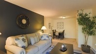 This single-floor space showcases an open-plan layout that seamlessly connects the living and dining areas, creating a cohesive and functional environment. The living area is anchored by a plush, neutral-toned sofa dressed with decorative pillows, positioned on a patterned area rug over wall-to-wall carpeting. A bold, dark accent wall serves as a striking focal point, adorned with an oversized decorative clock that adds character and visual depth to the room. A table lamp provides warm, ambient lighting, complementing the overhead flush-mount ceiling light. The adjoining dining area transitions naturally from the living space and features a dark wood dining table surrounded by matching chairs, topped with tasteful table décor. Decorative wall art accents this area, adding a finishing touch to the overall aesthetic. The warm, neutral color palette throughout — highlighted by the contrast of the dark accent wall — creates an inviting and balanced ambiance. The layout is practical and well-proportioned, making efficient use of the available space while maintaining a comfortable and welcoming atmosphere suitable for everyday living and entertaining.