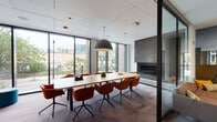 Located at 845 W Madison St in Chicago's West Loop neighborhood, this single-floor amenity space offers a thoughtfully designed environment that balances collaborative and private work settings. The layout incorporates distinct zones connected by consistent modern design language throughout.

The conference room is a standout feature, anchored by a sleek wood table surrounded by terracotta-hued swivel chairs and illuminated by a striking pendant light. Floor-to-ceiling windows frame views of the surrounding streetscape, flooding the room with natural light, while a linear fireplace set into a textured stone wall adds a refined touch. Glass partitions maintain visual connectivity with adjacent areas.

The open lounge area is defined by exposed concrete columns, a vertical wood slat screen partition, and a mix of high-back yellow lounge seating, gray sofas, and ottoman poufs arranged on patterned carpet. This zone benefits from abundant natural light from perimeter windows overlooking greenery.

A dedicated kitchenette features rich navy cabinetry, a three-dimensional tile backsplash, and warm wood flooring, bordered by black-framed glass walls. Additional glass-enclosed workrooms provide semi-private settings with casual lounge furnishings, a wall-mounted display, and views into the broader amenity floor, creating a flexible environment suited for both focused work and informal collaboration.