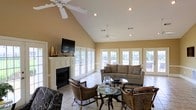 This single-floor space makes a striking impression with its dramatic vaulted ceilings, which lend a sense of openness and grandeur to the main living area. Dual ceiling fans are thoughtfully positioned throughout, ensuring comfortable airflow beneath the high, angled roofline. The focal point of the room is a classic white-trimmed fireplace, above which a wall-mounted television is installed, seamlessly blending traditional charm with modern convenience. Decorative accents on the mantel add a refined touch to the space.

Natural light is a defining feature here, with an expansive arrangement of white-framed French doors lining the exterior walls. These doors provide sweeping views of the lush outdoor surroundings and create a seamless connection between the interior and the outside. The warm, golden-toned walls complement the light tile flooring, which runs throughout the space and contributes to the bright, airy atmosphere.

The living area is furnished with a comfortable neutral-toned sofa, wicker accent chairs, and glass-topped coffee tables, creating an inviting and casual seating arrangement. Recessed lighting supplements the abundant natural light, ensuring the space is well-lit at any time of day. Overall, the room exudes a relaxed, sun-filled ambiance ideal for both everyday living and entertaining.