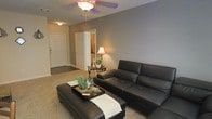 This single-floor residence offers a thoughtfully designed layout that balances comfort and functionality. The living area serves as the heart of the space, featuring neutral gray walls that create a calm, modern backdrop. A ceiling fan with a light fixture anchors the room from above, providing both airflow and ambient illumination. The space is furnished with a sleek black leather sectional sofa paired with a matching ottoman that doubles as a coffee table, styled with a decorative tray, greenery, and accent pieces that add a touch of personality. Warm lighting from a classic table lamp on a side accent table enhances the cozy atmosphere, complemented by a small potted plant that brings a touch of nature indoors. The living area flows naturally toward an entryway with a solid front door, offering a clear sense of arrival and transition. A doorway visible in the background hints at additional rooms beyond, suggesting a connected and practical floor plan. The carpeted flooring throughout adds warmth and comfort underfoot. Overall, this residence presents a clean, inviting aesthetic with a harmonious blend of neutral tones and contemporary furnishings that make it move-in ready.