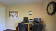 This single-floor workspace is designed as a functional office environment, offering a straightforward and practical setup for computer-based work. The room features warm, light yellow walls that create a neutral and approachable atmosphere, complemented by a textured ceiling and carpeted flooring.

At the heart of the space sits a shared desk running along one wall, accommodating two workstations side by side. Each station is equipped with a monitor and keyboard, with ergonomic black office chairs providing seating. A laser printer is centrally positioned between the two monitors, making it accessible to both users and supporting shared printing needs.

A cork bulletin board mounted on one wall serves as an informational hub, displaying notices, papers, and colorful lettering — suggesting active use in a communal or administrative setting. A framed document is also mounted on the wall above the desk area, adding an organized, professional touch.

The space is modest in scale but efficiently arranged to maximize functionality. Natural light appears limited, with the room relying primarily on interior lighting. Overall, the workspace conveys a utilitarian, productive environment suited for administrative or office support tasks.