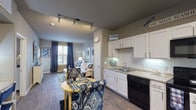 This single-story unit is designed with an open-concept layout that connects the kitchen and living areas into one cohesive space. The kitchen is outfitted with white cabinetry, light granite-style countertops, a white subway tile backsplash, and black appliances including a range, built-in microwave, dishwasher, and refrigerator. Track lighting overhead and under-cabinet lighting ensure the kitchen is well illuminated. A small dining area sits adjacent to the kitchen, providing a functional space for everyday meals.

Wood-look plank flooring extends throughout the main living areas, adding warmth and visual continuity. The living room receives natural light through a window dressed with navy curtains. Neutral gray walls serve as a consistent backdrop across the entire unit.

Two bedrooms feature plush carpeting, ceiling fans, and windows with blinds and decorative curtains. Each room offers comfortable proportions suitable for standard bedroom furnishings. The full bathroom includes a tub-shower combination with tile surround, a single-sink vanity with white cabinetry, and wood-look flooring. Directly accessible from the bathroom is a walk-in closet with shelving and hanging storage, adding practical organizational space to the unit.
