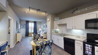 This single-story unit is designed with an open-concept layout that connects the kitchen and living areas into one cohesive space. The kitchen is outfitted with white cabinetry, light granite-style countertops, a white subway tile backsplash, and black appliances including a range, built-in microwave, dishwasher, and refrigerator. Track lighting overhead and under-cabinet lighting ensure the kitchen is well illuminated. A small dining area sits adjacent to the kitchen, providing a functional space for everyday meals.

Wood-look plank flooring extends throughout the main living areas, adding warmth and visual continuity. The living room receives natural light through a window dressed with navy curtains. Neutral gray walls serve as a consistent backdrop across the entire unit.

Two bedrooms feature plush carpeting, ceiling fans, and windows with blinds and decorative curtains. Each room offers comfortable proportions suitable for standard bedroom furnishings. The full bathroom includes a tub-shower combination with tile surround, a single-sink vanity with white cabinetry, and wood-look flooring. Directly accessible from the bathroom is a walk-in closet with shelving and hanging storage, adding practical organizational space to the unit.