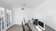 This single-floor office space offers a clean, functional environment designed to support focused, productive work. The interior is finished with crisp white walls and dark hardwood-style flooring, creating a sharp contrast that lends the space a professional and modern aesthetic.

At the heart of the office is a large, L-shaped wood-toned desk that comfortably accommodates two workstations, each equipped with an all-in-one desktop computer and keyboard. A multifunction printer sits centrally on the desk, making it easily accessible from both stations. Ergonomic office chairs complete the setup, ensuring comfort during extended work sessions.

Natural light is a defining feature of this space. A glass-paneled entry door and a set of elegant French doors flood the interior with sunlight and offer views of the surrounding greenery, creating an inviting and open atmosphere despite the office's compact footprint. The French doors also provide direct access to an adjacent area, adding versatility to the layout.

A textured ceiling, recessed HVAC vents, and a wall-mounted thermostat reflect thoughtful climate control throughout. Interior doors allow for privacy and separation as needed. Overall, this office space strikes an effective balance between practicality and comfort, making it well-suited for small teams or collaborative work environments.