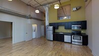 This single-floor loft showcases a striking industrial aesthetic, blending raw architectural elements with modern finishes to create a distinctive and functional living space. Soaring ceilings draw the eye upward to exposed ductwork, visible pipes, and structural columns that lend an authentic urban character throughout. Pendant lighting hangs from above, casting a warm glow across the open-concept floor plan. The kitchen is a standout feature, outfitted with dark navy cabinetry that contrasts boldly against a bold olive-green accent wall. Stainless steel appliances, including a full-size refrigerator, a gas range, and an over-the-range microwave, complement the dark countertops and undermount sink, delivering both style and practicality. Warm-toned hardwood flooring runs throughout the main living area, adding texture and continuity to the space. Large windows positioned high on the walls invite natural light deep into the interior. A dedicated laundry area with a stacked washer and dryer is neatly tucked behind a doorway, offering in-unit convenience. Additional doorways suggest private rooms or utility spaces beyond the main living area. Overall, the space exudes an open, airy, and thoughtfully designed industrial-modern ambiance.