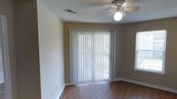 This single-story space offers a clean and functional layout designed for comfortable everyday living. The main living area is highlighted by warm wood-look hardwood flooring that runs throughout, providing a welcoming and cohesive aesthetic. A ceiling fan with a built-in light fixture is centrally mounted, ensuring both air circulation and ambient lighting. The textured popcorn ceiling adds a classic residential character to the space. Natural light is a key feature, with a large sliding glass door dressed in vertical blinds serving as the focal point of the room. The door opens to an outdoor area, allowing for seamless indoor-outdoor flow and views of the surrounding greenery. An additional window on the adjacent wall further enhances the brightness of the space. Neutral-toned walls complement the flooring and provide a versatile backdrop for any furnishing style. White baseboards frame the room with a crisp, clean finish. The overall ambiance is practical and inviting, with simple, timeless design elements that create a bright and airy environment suitable for a variety of uses.