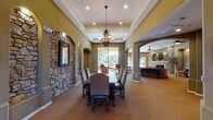 This single-story community clubhouse showcases a warm Tuscan-inspired aesthetic throughout its thoughtfully designed gathering spaces. The grand entrance welcomes visitors with natural stone tile flooring, double glass-paneled doors, and an adjacent leasing or administrative office, setting an inviting tone from the moment of arrival.

The formal dining area serves as a central highlight, featuring a coffered tray ceiling, an ornate wrought-iron chandelier, and a long wooden dining table surrounded by upholstered chairs. Dramatic stone-clad arched niches line the walls, adding rich architectural texture and depth. Arched passageways connect this space seamlessly to adjacent areas.

The lounge area offers a comfortable retreat with vaulted ceilings, a ceiling fan, plush seating, and a stone-surround fireplace with an arched decorative alcove. Large windows and a glass door provide direct views of and access to an outdoor pool area, filling the room with natural light.

A well-appointed kitchen features rich cherry wood cabinetry, granite countertops, a tile backsplash, and black appliances including a dishwasher and microwave, making it ideal for catered events or community gatherings.

Adjacent to the lounge, a game room with a billiard table and its own stone fireplace rounds out the amenities. Warm olive and earth-toned walls flow consistently throughout, creating a cohesive, welcoming atmosphere.