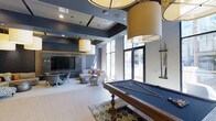 This single-floor amenity lounge is a well-appointed social space designed for both entertainment and relaxation. At its centerpiece sits a classic wooden billiards table with a slate-blue felt surface, positioned atop a decorative area rug and illuminated by a series of large, warm-toned drum pendant lights that hang from the elevated ceiling. Cue sticks are neatly mounted on a wall-mounted rack nearby, adding a functional yet stylish touch.

The space transitions seamlessly into a comfortable lounge area, where a curated arrangement of plush sofas and accent chairs surrounds a coffee table, creating an inviting setting for casual conversation. A large flat-screen television is mounted against a textured feature wall, complemented by subtle ambient lighting that enhances the room's sophisticated atmosphere.

Floor-to-ceiling windows and glass doors line one wall, flooding the interior with abundant natural light while offering views of an outdoor area, likely a pool or courtyard. The overall design palette features cool grays and blues accented by warm lighting, contributing to a modern, upscale ambiance throughout.