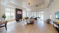 Located at 386 Attenborough Dr in Rosedale, MD, this single-floor community clubhouse is thoughtfully designed to encourage social interaction and relaxation. The open-concept layout is anchored by a spacious lounge area featuring a dramatic floor-to-ceiling dark slate fireplace with a mounted flat-screen TV, creating a warm and inviting focal point. Soaring ceilings, recessed lighting, and a stylish clustered pendant fixture add architectural interest throughout the space. Light wood-tone flooring runs seamlessly across the entire floor, contributing to a bright and cohesive aesthetic. Large, multi-pane windows flood the interior with abundant natural light, while also offering direct views of the outdoor pool area — accessible via glass-paned doors. The dining and café-style seating zone features high-top tables and modern chairs arranged near the window-lined perimeter, ideal for casual gatherings or work. A secondary lounge nook includes a wall-mounted TV, console table, and intimate bistro seating. Tasteful greenery, including potted fiddle-leaf fig trees, adds life and warmth throughout. The community's ethos is proudly displayed with the wall phrase "Building Community. Building Memories." — setting the tone for a welcoming, people-centered environment.