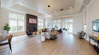 Located at 386 Attenborough Dr in Rosedale, MD, this single-floor community clubhouse is thoughtfully designed to encourage social interaction and relaxation. The open-concept layout is anchored by a spacious lounge area featuring a dramatic floor-to-ceiling dark slate fireplace with a mounted flat-screen TV, creating a warm and inviting focal point. Soaring ceilings, recessed lighting, and a stylish clustered pendant fixture add architectural interest throughout the space. Light wood-tone flooring runs seamlessly across the entire floor, contributing to a bright and cohesive aesthetic. Large, multi-pane windows flood the interior with abundant natural light, while also offering direct views of the outdoor pool area — accessible via glass-paned doors. The dining and café-style seating zone features high-top tables and modern chairs arranged near the window-lined perimeter, ideal for casual gatherings or work. A secondary lounge nook includes a wall-mounted TV, console table, and intimate bistro seating. Tasteful greenery, including potted fiddle-leaf fig trees, adds life and warmth throughout. The community's ethos is proudly displayed with the wall phrase "Building Community. Building Memories." — setting the tone for a welcoming, people-centered environment.