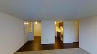 This single-floor residence offers a straightforward and functional layout designed for comfortable everyday living. The main living area is open and airy, featuring dark hardwood flooring that extends throughout the space, providing a warm contrast against the crisp white walls and smooth ceilings. The open-concept design allows the living area to flow naturally into adjacent spaces, creating a cohesive and connected feel.

The kitchen is accessible directly from the main living area and is outfitted with medium-toned wood cabinetry, a white refrigerator, a gas range, and a sink positioned beneath under-cabinet lighting. The warm ambient lighting in the kitchen creates an inviting atmosphere that complements the rest of the home.

A secondary room, visible through a wide doorway, features the same hardwood flooring and white walls, offering flexible use as a bedroom or additional living space. A closet with louvered doors adds practical storage, while a front entry door with hardware completes the layout. The space maintains a clean, neutral aesthetic throughout, making it easy to personalize and furnish to suit a variety of lifestyles.