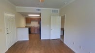 This single-story space presents an open-concept design that seamlessly connects the living area to the kitchen. Hardwood flooring extends throughout the main living space, adding warmth to the neutral, beige-toned walls accented by crown molding. The kitchen is equipped with medium-toned wooden cabinetry, a stainless steel refrigerator, a dishwasher, and a sink with granite-style countertops. A peninsula counter separates the kitchen from the living area, offering additional workspace and a natural division between the two zones. Overhead fluorescent lighting illuminates the kitchen, while a ceiling fan services the living area. HVAC vents are positioned throughout the ceiling for consistent climate control. A doorway visible from the main living space leads to an additional room, partially visible and featuring built-in counter space, suggesting a dedicated workspace or secondary functional area. The layout is straightforward and practical, with a clean, understated aesthetic throughout. The overall ambiance is modest and functional, well-suited for everyday living with an efficient use of the available space.