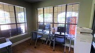 This single-story space functions as a shared business center or computer lab, designed to provide a productive and accessible environment for its users. The room is equipped with multiple workstations, each featuring a desktop computer, monitor, keyboard, and rolling ergonomic chair, arranged along sleek, minimalist desks positioned against the far wall.

One of the most notable features of this space is the expansive grid-pane window wall, which spans nearly the full width and height of the room, allowing abundant natural light to flood the interior and offering views of an outdoor courtyard area with mature trees and neighboring brick architecture. A secondary window on the adjacent wall further enhances the brightness of the room.

The flooring consists of warm hardwood-style planks, complementing the olive-green painted walls to create a welcoming yet professional atmosphere. White baseboards provide a clean contrast throughout. A Coca-Cola vending machine is visible through the window, suggesting proximity to a communal amenity area. The overall layout is practical and organized, making efficient use of the available space.