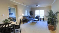 This single-floor space features an open-concept living and dining area finished with soft, neutral-toned carpeting throughout. The living room is anchored by a blue upholstered sofa and matching armchair, arranged around a dark wood coffee table. Crown molding lines the ceiling, adding a refined architectural detail, while a ceiling fan with a light fixture provides both ventilation and overhead illumination. Large windows along one wall allow natural light to fill the space, contributing to an airy, welcoming ambiance. A framed piece of artwork and a decorative potted plant add character to the room. A floor lamp provides warm accent lighting near the seating area. To one side, a small dining set with dark wood chairs is partially visible, offering a dedicated area for meals. A doorway visible from the living area leads to what appears to be a bedroom, suggesting a well-connected layout. The neutral wall tones and consistent flooring create a cohesive aesthetic throughout, making the space feel unified and functional for everyday living.