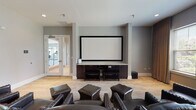 Located at 13976 John Humphrey Dr in Orland Park, Illinois, this single-floor screening and media room is designed to deliver a refined entertainment experience. The focal point of the space is a large, wall-mounted projection screen set against a recessed accent wall, complemented by a dark wood media console housing audio-visual equipment and a subwoofer. Rows of dark leather armchairs with nailhead trim detailing and matching ottomans are arranged theater-style, providing comfortable seating for multiple viewers. The flooring throughout is light-toned hardwood, adding warmth and continuity to the space. A large bank of windows along one wall is dressed with floor-length draping curtains, allowing natural light to filter in while offering views of the surrounding greenery and neighborhood. Recessed and ceiling-mounted lighting provides adjustable illumination for varied uses. A ceiling-mounted projector is visible overhead, reinforcing the room's dedicated media function. A set of glass-panel French doors connects the screening room to an adjacent fitness area, visible through the doors with exercise equipment inside. The neutral gray and white color palette throughout lends a clean, contemporary feel, making the space well-suited for both private screenings and communal gatherings.