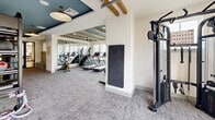 Located at 3700 Limestone Parkway in Gainesville, Georgia, this fitness center offers a single-floor exercise room thoughtfully designed to support a comprehensive workout experience. The space features a distinctive aesthetic, with tray ceilings painted in a deep teal-blue accented by exposed rustic wood beams, ceiling fans, and pendant lighting that together create an inviting yet energizing atmosphere. Neutral-toned carpet flooring extends throughout, providing comfort underfoot across the generously sized workout area. Large windows along multiple walls flood the interior with natural light while offering views of the surrounding landscape.

The cardio zone is outfitted with treadmills, ellipticals, stair climbers, a rowing machine, and stationary bikes. The strength training section includes a TRX suspension rig, cable machines, weight benches, barbells, dumbbells, kettlebells, medicine balls, and battle ropes, catering to a wide range of training modalities. Full-length mirrors line sections of the walls, enhancing the sense of space and allowing users to monitor their form. A dedicated area for free weights and functional accessories is clearly organized with racks and storage. A garage-style door opens to an adjacent outdoor turf area, seamlessly connecting the interior to an exterior training space ideal for functional fitness activities.