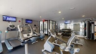 This fitness center is a comprehensively outfitted facility designed to support a full range of workout routines across a single, open floor plan. The space is divided into two primary functional zones that flow seamlessly into one another. The cardio area is anchored by large floor-to-ceiling windows framed in sleek black metal, flooding the zone with natural light. Equipment here includes treadmills, elliptical trainers, stationary bikes, and a rowing machine, all positioned to take advantage of the bright, airy atmosphere. Wall-mounted flat-screen TVs are strategically placed throughout for entertainment during workouts. The strength training zone features a bold accent wall in warm amber-orange, creating a high-energy visual contrast to the cooler gray tones elsewhere. This area is stocked with a full rack of dumbbells in graduated weights, a multi-functional cable machine, a Cybex squat rack, a weight bench, and a collection of medicine balls. Mirrored walls expand the sense of space and allow for form monitoring. The flooring throughout is durable rubber gym matting, and recessed lighting supplements the natural light. A glass-enclosed vestibule serves as an entry passage, adding a polished, modern touch to the overall design.