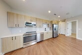 This single-floor unit showcases a thoughtfully designed layout with a clean, contemporary aesthetic throughout. The kitchen is a standout feature, equipped with light wood flat-front cabinetry accented by matte black hardware, white quartz countertops, and a white tile backsplash. A full suite of stainless steel appliances includes a range, built-in microwave, dishwasher, and a side-by-side refrigerator with an in-door ice and water dispenser. Light wood-style luxury vinyl plank flooring extends through the kitchen and into the open living and dining area, which benefits from multiple large windows fitted with horizontal blinds, allowing for generous natural light and outdoor views. A door provides direct access to an outdoor area from the living space. The hallway connects the main living areas to the private rooms, featuring recessed lighting and additional doors to closets or utility spaces. Bedrooms are finished with plush carpeting, ceiling fans, and wide windows that invite ample daylight. The bathroom offers a wide vanity with light wood cabinetry, white quartz countertop, an undermount sink with a modern faucet, and a large backlit mirror, all set on wood-style flooring.