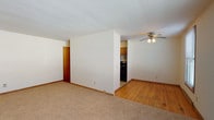 This single-floor residence offers a practical and comfortable layout with a thoughtful mix of flooring materials and functional design throughout. The living area is spacious and bright, featuring warm beige carpeting, white walls with clean baseboard trim, and a ceiling fan with light fixture that enhances air circulation and illumination. Natural light filters in through a large window, adding an open, airy quality to the space. The kitchen is efficiently arranged with warm honey-toned wood cabinetry, granite-patterned countertops, a stainless steel sink, and black appliances including a full-size refrigerator. Tile flooring in the kitchen provides a clean and practical surface. The bedrooms are well-proportioned, showcasing gleaming hardwood floors that add warmth and character. One bedroom features dual windows that invite abundant natural light, a ceiling fan, and direct outdoor views. Another bedroom includes an open closet with a dedicated storage rod and additional window. The bathroom is clean and straightforward, equipped with a single-sink vanity featuring wood cabinetry, tile flooring, and a full tub-shower combination with tile surround. Overall, this residence presents a neat, move-in-ready environment with consistent finishes and a cohesive, functional flow throughout its rooms.