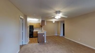This single-floor residence offers a practical and comfortable open-concept layout that seamlessly connects the kitchen and living area. The kitchen is equipped with black appliances, including a double oven and range, complemented by warm honey-toned wood cabinetry and a peninsula with a sink that doubles as a convenient breakfast bar. Hard flooring in the kitchen transitions smoothly into plush wall-to-wall carpeting in the main living area, creating a cozy and inviting atmosphere. The living space is generously proportioned, providing ample room for a variety of furniture arrangements. A ceiling fan with an integrated light fixture is centrally positioned overhead, contributing to both comfort and ambient lighting. Warm beige walls and white baseboards run throughout, lending the space a clean, neutral palette that is easy to personalize. A textured ceiling adds subtle visual character. A hallway visible from the main living area leads to additional rooms, suggesting a well-organized and functional floor plan. Overall, the space delivers a welcoming, move-in-ready environment with a balanced blend of practicality and comfort.