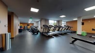 This fitness center is a comprehensively outfitted facility designed to accommodate a wide range of workout needs across a single, expansive floor. The space is organized into distinct functional zones, each dedicated to a specific type of exercise, creating an efficient and practical layout for users.

The cardio section is prominently featured, housing multiple treadmills with integrated display screens, stationary cycling bikes, and elliptical machines arranged in rows along the windowed perimeter. Large windows along the exterior walls allow natural light to fill the space and provide views of the surrounding area, enhancing the workout environment.

A dedicated strength training zone includes a full power rack with barbell and weight plates, multiple weight benches, and an organized dumbbell rack housed within built-in white shelving units. Wall-mounted mirrors in this area support safe and effective training. Adjacent to this, a selection of cable and resistance machines, accompanied by wall-mounted televisions, form a sizable machine training area set against warm amber-toned accent walls.

Additional amenities include built-in storage cabinetry, water fountains, and a rowing machine for varied cardio options. The flooring throughout is dark, durable rubber, well-suited for heavy equipment use. Recessed and flush-panel ceiling lighting ensures consistent illumination across all zones, contributing to a clean and functional atmosphere.