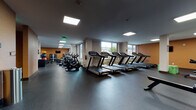 This fitness center is a comprehensively outfitted facility designed to accommodate a wide range of workout needs across a single, expansive floor. The space is organized into distinct functional zones, each dedicated to a specific type of exercise, creating an efficient and practical layout for users.

The cardio section is prominently featured, housing multiple treadmills with integrated display screens, stationary cycling bikes, and elliptical machines arranged in rows along the windowed perimeter. Large windows along the exterior walls allow natural light to fill the space and provide views of the surrounding area, enhancing the workout environment.

A dedicated strength training zone includes a full power rack with barbell and weight plates, multiple weight benches, and an organized dumbbell rack housed within built-in white shelving units. Wall-mounted mirrors in this area support safe and effective training. Adjacent to this, a selection of cable and resistance machines, accompanied by wall-mounted televisions, form a sizable machine training area set against warm amber-toned accent walls.

Additional amenities include built-in storage cabinetry, water fountains, and a rowing machine for varied cardio options. The flooring throughout is dark, durable rubber, well-suited for heavy equipment use. Recessed and flush-panel ceiling lighting ensures consistent illumination across all zones, contributing to a clean and functional atmosphere.