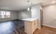 This single-floor unit offers a thoughtfully designed open-concept layout that seamlessly connects the kitchen and living area. The kitchen is a standout feature, equipped with stainless steel appliances including a smooth-top electric range, over-the-range microwave, and a refrigerator. White cabinetry with sleek hardware contrasts beautifully with a white quartz countertop, while a classic white subway tile backsplash adds a clean, modern touch. A central island with light wood cabinetry provides additional prep and storage space, illuminated by pendant lighting overhead. Recessed lighting throughout enhances the bright, airy atmosphere.

The open living area is generously proportioned, featuring warm wood-tone laminate flooring that runs throughout the unit, neutral taupe-toned walls, and clean white baseboards. Natural light filters in through windows that offer views of the surrounding urban greenery.

The bedroom is a comfortable, well-sized room with the same wood-tone flooring and access to a closet. The bathroom is finished in light neutral tones, featuring a full soaking tub with a shower, a single-sink vanity with white cabinetry, and tile flooring. Overall, the unit presents a clean, contemporary aesthetic with functional, modern finishes.