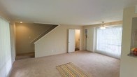 This two-story home features a spacious main living area finished with plush wall-to-wall carpeting and neutral beige tones throughout, creating a warm and inviting atmosphere. A staircase along one wall leads to the upper floor, with the underside adding architectural interest to the room. A ceiling fan is centrally positioned, providing both lighting and air circulation. Built-in shelving beside a large window offers practical storage and display space, while vertical blinds on the window allow for adjustable natural light. A doorway visible from the main living area opens to an adjacent room or hallway, suggesting a connected and functional floor plan. The overall design is clean and understated, emphasizing openness and practicality. The neutral palette and carpeted flooring throughout contribute to a comfortable, cohesive aesthetic suited for everyday living.