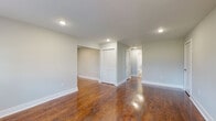 This single-floor space offers a clean, modern aesthetic defined by rich, warm-toned hardwood flooring that runs throughout the main living areas and bedrooms. The neutral gray walls create a bright, airy atmosphere that complements the polished floors and white trim details.

The open living area is illuminated by recessed lighting and benefits from natural light filtering through windows fitted with blinds. A louvered closet provides convenient built-in storage. The layout flows seamlessly through a hallway connecting the main rooms, as seen in the overhead dollhouse view.

The kitchen is outfitted with dark-stained cabinetry, crisp white countertops, and a classic white subway tile backsplash. Stainless steel appliances — including a gas range and over-the-range microwave — give the kitchen a contemporary, functional character. The flooring transitions to a light tile in this area, adding visual distinction.

The bedrooms continue the hardwood flooring theme, with one room featuring a double window with baseboard heating beneath it, offering a warm, well-lit retreat. The bathroom is a standout feature, finished with striking marble-patterned wall tile surrounding a tub-shower combination with a sliding glass door, complemented by neutral floor tiles and a window for natural ventilation. Overall, the space presents a cohesive, refreshed interior with quality finishes throughout.