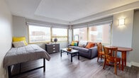 This studio in Ann Arbor, Michigan, offers a well-designed, single-floor layout that efficiently combines living, sleeping, and dining functions within a compact footprint. The main living area is bright and open, anchored by a bank of large windows with gold-trimmed frames that flood the space with natural light and offer views of the surrounding Ann Arbor cityscape. Wood-toned luxury vinyl plank flooring runs throughout, lending a clean and cohesive aesthetic.

The sleeping and living areas share one open space, allowing for flexible furniture arrangement. A dedicated dining nook sits adjacent to the kitchenette, which features white cabinetry, a subway tile backsplash, stainless steel appliances including a gas range, refrigerator, and microwave, as well as a single-basin sink with ample counter space. Wall-mounted lighting in the kitchen area adds both function and a modern touch.

The bathroom is neatly appointed with a white vanity featuring a light stone countertop, built-in open shelving, and a full tub-shower combination with a curtain rod and grab bar. Neutral gray tones throughout the bathroom contribute to a calm, clean atmosphere.

A closet with louvered doors provides additional storage. The textured ceiling, clean white walls, and consistent flooring tie the entire space together into a practical and inviting urban studio.