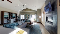 This single-floor space makes a strong impression with its dramatic vaulted ceiling, which rises to a prominent peak and is complemented by a ceiling fan and recessed lighting that together create a bright, airy atmosphere. The open-concept living area is generously proportioned, allowing natural light to pour in through multiple windows dressed in sheer, patterned curtains. The living room centers around a large sectional sofa in a neutral gray, adorned with teal and patterned throw pillows that coordinate with the bold teal area rug beneath a round glass coffee table. A floor-to-ceiling tiled fireplace with a mounted television serves as a striking focal point on one wall, flanked by a modern wall sconce. Vibrant artwork, including a large expressive portrait, adds a creative energy to the space. Toward the rear, an open dining or workspace area is visible, featuring bar-height seating and additional natural light from surrounding windows. The light wood-toned flooring flows throughout, tying the spaces together cohesively. The overall ambiance is contemporary and welcoming, balancing comfort with stylish, curated design elements.