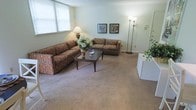 This single-floor space offers a warm and functional layout designed for comfortable everyday living. The living room serves as the central gathering area, featuring wall-to-wall carpeting that adds a sense of coziness underfoot. Two patterned sofas are arranged around a dark wood coffee table, creating an inviting seating arrangement well-suited for relaxation or casual conversation. Natural light flows generously into the room through large windows fitted with horizontal blinds, brightening the neutral-toned walls and giving the space an open, airy feel. A tall floor lamp and a classic table lamp provide layered ambient lighting for evening use. Framed artwork adorns the walls, adding a touch of personality to the otherwise understated décor. A decorative floral arrangement on the coffee table and a potted plant in the corner introduce natural elements that soften the interior. The layout provides ample open floor space, contributing to a sense of roominess and ease of movement throughout. Overall, the space presents a clean, traditional aesthetic with practical furnishings and a comfortable, livable ambiance suitable for a range of occupants.