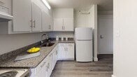 This single-floor unit presents a clean, modern aesthetic defined by neutral tones and consistent hardwood-style flooring that runs throughout every room. The living areas are bright and open, featuring light gray walls, crisp white baseboards, and windows that invite natural light while offering views of the surrounding neighborhood. Baseboard heating units are present in multiple rooms, ensuring year-round comfort. The kitchen is thoughtfully equipped with white shaker-style cabinetry fitted with brushed nickel hardware, granite-style countertops, a stainless steel sink with a modern faucet, and a full-size refrigerator. Under-cabinet lighting enhances both functionality and ambiance in the kitchen space. The bathroom is neatly appointed with a white vanity, a framed mirror, a tub-shower combination with tiled surround, and a bold patterned shower curtain that adds a touch of character. A dedicated utility closet houses the water heater, keeping mechanical systems organized and out of the way. Throughout the unit, the design emphasizes simplicity and practicality, with a neutral color palette and clean finishes that provide a versatile, move-in-ready environment.