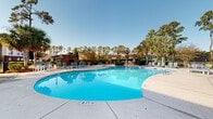 Located at 900 Brookstone Way in Goose Creek, South Carolina, this community amenity area centers around a generously sized freeform swimming pool with crystal-clear blue water, designed for both relaxation and recreation. The pool features multiple entry points and is surrounded by a broad concrete sundeck, furnished with lounge chairs and seating areas that invite residents to unwind under the South Carolina sun.

A charming brick outdoor fireplace anchors one end of the pool deck, providing a warm gathering spot during cooler evenings. A pergola structure at the far end of the pool area adds architectural character and additional shaded seating. Lush mature landscaping — including tall pine trees and swaying palm trees — frames the entire space, offering natural privacy and a distinctly coastal Southern atmosphere.

Adjacent to the pool is a well-maintained utility and facilities building constructed with a classic combination of white siding and brick detailing, housing pool equipment and support amenities. A gated entry provides controlled access to the pool area. The overall design reflects a welcoming, resort-inspired aesthetic that complements the surrounding residential community, making it a standout shared space for those living at Brookstone Way.