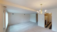 This single-floor space presents an open, airy layout finished in a clean, neutral palette throughout. The main living area is expansive, with smooth white walls and light beige carpeting that extends across the floor, creating a bright and cohesive atmosphere. A structural beam runs along the ceiling, subtly defining zones within the open plan without disrupting the sense of spaciousness.

A standout feature of the space is the elegant gold candelabra-style chandelier, which serves as a refined focal point and adds a classic decorative touch to the otherwise understated interior. The ceiling is standard height and well-maintained, complementing the clean lines of the room.

Recessed HVAC vents are integrated into the walls, ensuring climate control while maintaining a tidy aesthetic. Multiple electrical outlets are distributed along the walls, providing practical utility throughout the space. An open passageway at the far end of the room connects to an adjacent area, suggesting additional rooms or functional zones beyond the main living space. Overall, the space offers a blank canvas with solid foundational finishes, well-suited for customization.