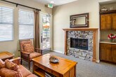 The Bungalows at Chapman Heights Apartment Homes, located in Yucaipa, California, presents a thoughtfully designed single-story community clubhouse that serves as a central gathering space for residents. The entryway greets visitors with elegant tile flooring accented by a decorative inlay border, soaring ceilings, a statement chandelier, and rich warm-toned wall colors that establish a welcoming craftsman-inspired aesthetic throughout. Built-in wood cabinetry with display shelving lines the entry hall, complemented by wall sconces and framed floor plan displays beneath The Bungalows signage. The main lounge area transitions to plush carpeting and features a striking stacked-stone fireplace with a classic wood mantel and a framed mirror above, flanked by additional built-in cabinetry with countertop workspace. Comfortable upholstered seating, a wood coffee table, and large windows with drape panels allow abundant natural light to flow in, while a glass door provides direct access to an outdoor patio area. A dedicated fitness room is equipped with a multi-station strength training unit, a recumbent exercise bike, wall-mounted television, and carpeted flooring — offering residents a convenient on-site workout space. The overall ambiance is warm, functional, and community-focused, blending craftsman design details with practical amenities.