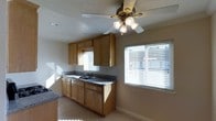 This single-story space showcases a well-appointed kitchen that serves as a central feature of the layout. The kitchen is outfitted with warm honey-toned wood cabinetry, offering generous upper and lower storage options. Dark granite countertops provide a contrasting, durable work surface, complemented by a double-basin sink positioned beneath a window. The light tile flooring throughout contributes to a clean, cohesive aesthetic. Natural light is a notable feature, with multiple windows dressed in horizontal blinds allowing sunlight to fill the space throughout the day. A ceiling fan with an integrated light fixture is centrally mounted, enhancing air circulation and providing additional illumination. The neutral wall tones and simple trim details create a straightforward, functional ambiance. The overall design reflects a practical, traditional style with classic finishes that offer a comfortable and utilitarian environment suitable for everyday living.