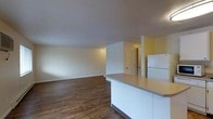 This single-floor residence offers a clean and functional open-concept design that seamlessly connects the kitchen and living areas into one cohesive space. The kitchen is anchored by a white cabinetry base with a curved peninsula counter finished in a neutral laminate surface, providing both prep space and a natural division between the kitchen and the expansive living area. A white refrigerator stands adjacent to the peninsula, completing the kitchen's practical layout. Rich hardwood-style flooring runs throughout the main living space, adding warmth and visual continuity to the interior. The walls are painted in soft, neutral tones — a warm yellow accent wall adds a touch of color near the entryway corridor, while crisp white walls define the living area. The layout feels open and airy, with a spacious living zone that offers flexibility for various furniture arrangements. A paneled interior door visible in the background leads to additional rooms, suggesting a well-organized floor plan. The smooth ceilings and clean lines throughout the space contribute to a simple, uncluttered ambiance that is both welcoming and easy to personalize.