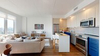 This contemporary single-floor space is thoughtfully designed with an open-concept layout that seamlessly connects the kitchenette, dining, and living areas. The kitchenette is a standout feature, set against a bold teal accent wall adorned with vibrant blue mosaic tile backsplash. White cabinetry, stainless steel appliances, and a sleek countertop complete the functional yet stylish setup. A striking gold Sputnik pendant light anchors the dining area, where a large white table surrounded by mixed seating — including woven rattan and wood-seated chairs — creates an inviting gathering space. Large-format light stone tile flooring runs throughout, lending the space a clean, cohesive feel. The living area is bathed in natural light, courtesy of expansive floor-to-ceiling windows framed in dark metal. Sheer white curtains soften the light while offering sweeping outdoor views, including a cityscape visible from one vantage point and lush greenery from another. A generous sectional sofa in a neutral tone anchors the lounge space, which also features a wall-mounted TV set against warm wood paneling. The overall ambiance is modern, vibrant, and welcoming — a well-curated blend of bold color accents and clean contemporary design.