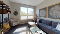 Located at 301–373 23rd St in Oakland, CA, this modern single-floor unit offers a thoughtfully designed open-concept layout. The kitchen is equipped with stainless steel appliances, including an electric range, microwave, dishwasher, and refrigerator, all complemented by crisp white cabinetry with dark countertops and under-cabinet lighting. A freestanding dining table with bar-height seating sits adjacent to the kitchen, creating a seamless transition between cooking and dining spaces. The living area receives generous natural light through large windows overlooking tree-lined urban views of Oakland. The bedroom is a calm, well-proportioned retreat featuring large windows that bring in abundant daylight and views of the surrounding cityscape, a walk-in closet, and plush carpeting. The bathroom features a vanity with a dark countertop, a wide mirror with overhead lighting, and a clean, minimal aesthetic with light-toned cabinetry. Throughout the unit, wood-style flooring anchors the main living areas, while neutral warm-gray wall tones create a cohesive and contemporary atmosphere. Recessed lighting throughout enhances the clean, modern feel of this well-appointed Oakland residence.