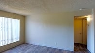 This single-story space presents a straightforward and functional layout with a clean, neutral aesthetic. The room showcases durable hardwood-style flooring in a warm, medium-toned wood finish, providing a grounded and practical foundation throughout. The walls are finished in a soft, muted beige-green tone, creating a calm and understated atmosphere that adapts easily to a variety of interior design styles. White baseboards provide a crisp contrast along the perimeter, lending a tidy, finished appearance to the room. The ceiling features a classic textured popcorn finish, a common characteristic of its era of construction. Electrical outlets are positioned along the walls at standard intervals, ensuring practical usability throughout the space. The room is open and unobstructed, offering flexibility in how it can be arranged and utilized. The overall ambiance is simple and neutral, making it an ideal blank canvas for those looking to personalize the space to their own taste. The combination of warm flooring tones and muted wall colors contributes to a quiet, livable environment well-suited for everyday use.