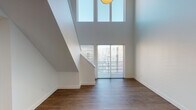 This two-story residence is defined by its striking double-height living area, where soaring ceilings draw the eye upward toward expansive clerestory windows that flood the space with natural light. A sliding glass door at the base of the living area frames city views, while a clean white staircase leads to the upper level. The main floor features hardwood-style flooring throughout the living and kitchen areas. The kitchen is equipped with dark espresso cabinetry reaching toward the tall ceiling, granite-style countertops, stainless steel appliances including a refrigerator, range, and built-in microwave, and a peninsula with pendant lighting above. A bedroom on the main level is carpeted and features a teal accent wall with a window overlooking the surrounding neighborhood. Its adjacent full bathroom includes a dark wood vanity with a granite-style countertop and a tub-shower combination. The upper level offers an open loft-style space with hardwood-style flooring, multiple windows with city views, and an open railing overlooking the living area below. A second full bathroom on this level features a granite vanity and a glass-enclosed shower.