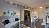 This single-floor loft-style space combines industrial design with modern comfort in a thoughtfully arranged open-concept layout. Exposed metallic ductwork runs prominently along the ceiling, serving as a defining architectural feature that adds an urban, industrial character to the interior. The polished concrete floors throughout create a sleek, cohesive foundation that reflects light and enhances the sense of spaciousness.

The kitchen is a central highlight, featuring dark cabinetry, stainless steel appliances including a refrigerator, range, and microwave, and a peninsula-style breakfast bar with modern pendant lighting suspended above. The bar seating with contemporary white stools creates a casual dining and gathering area that connects seamlessly to the living space.

The living area is bright and welcoming, furnished with a white sofa accented by navy throw pillows and complemented by a glass coffee table. Colorful abstract artwork on the walls adds a vibrant, artistic touch to the neutral palette. A warm table lamp provides soft ambient lighting.

Overall, the space strikes a balance between raw industrial aesthetics and refined modern living, making it a visually dynamic and functional environment well-suited for contemporary urban lifestyles.