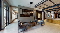 The Kilby amenity floor is a thoughtfully designed communal space that blends contemporary style with functional comfort. The entry lobby greets visitors with large floor-to-ceiling windows that invite abundant natural light, light-toned tile flooring, and a striking geometric pendant chandelier suspended above a curated seating arrangement of structured armchairs and a round wood-and-brass coffee table. A prominent illuminated "Kilby" sign anchors the feature wall, setting the tone for the space.

Moving deeper into the floor, an expansive lounge area opens up, distinguished by wide-plank pale wood flooring, dark wood-clad accent walls, and exposed beamed ceilings. Multiple seating vignettes with modern chairs, ottomans, and low tables are arranged around decorative fireplaces and curated artwork, creating an inviting atmosphere for socializing or relaxing.

A dedicated bar and kitchen area features bold emerald green cabinetry, quartz countertops, a draft beer tap, stainless steel appliances, and hexagonal tile flooring, complemented by a marble-topped bar with stylish swivel seating. A separate premium beverage station showcases dark wood cabinetry with brass hardware. The floor also includes a secure package locker room with smart lockers for convenient parcel retrieval.