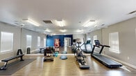Located at 810 E Slaughter Ln in Austin, Texas, this fitness center offers a well-equipped, single-floor exercise space designed to support a variety of workout routines. The room features durable hardwood-style flooring throughout, providing a clean and functional surface ideal for both equipment use and floor exercises.

The space is outfitted with a selection of cardio machines, including a treadmill, elliptical trainer, and stationary bike, as well as a rack of dumbbells, kettlebells, a weight bench, resistance tools, and balance equipment such as a BOSU ball and step platform. Full-length mirrors line one wall, enhancing the sense of openness and allowing users to monitor their form during workouts.

Multiple ceiling fans and bright fluorescent lighting ensure a comfortable, well-ventilated environment. Several windows fitted with blinds allow natural light to filter in while maintaining privacy. A bold blue accent wall adds a dynamic visual element to the otherwise neutral gray color scheme.

Convenience amenities include a beverage vending machine and a small under-counter refrigerator, making refreshments easily accessible. French-style doors provide entry and exit points. The overall layout is open and spacious, allowing for a variety of exercises without feeling crowded, making this facility a practical and functional resource for residents.