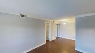 This single-floor space presents a clean, understated aesthetic defined by warm hardwood flooring and soft, neutral gray walls that create a calm, versatile backdrop. Crown molding runs along the ceiling perimeter, adding a touch of classic architectural detail to the otherwise straightforward design. The main living area is generously proportioned, offering an open feel well-suited for a variety of furniture arrangements.

A mirrored sliding closet provides both practical storage and the visual effect of expanded space, reflecting light throughout the room. An elegant chandelier anchors the far end of the room, contributing warmth and a refined finishing touch to the ceiling.

A doorway visible from the main area leads to an adjacent space, likely a bathroom or utility room, identifiable by its louvered door and interior shelving. Central air vents integrated into the ceiling and walls indicate a full HVAC system throughout.

White baseboards and trim provide crisp contrast against the darker flooring, reinforcing the space's tidy, well-maintained character. Overall, the space offers a bright, neutral canvas ready for personalization.